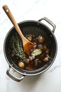 This image shows a soup pot being filled with caramelized onions, thyme, a bay leaf, and broth, combining ingredients for a hearty soup.