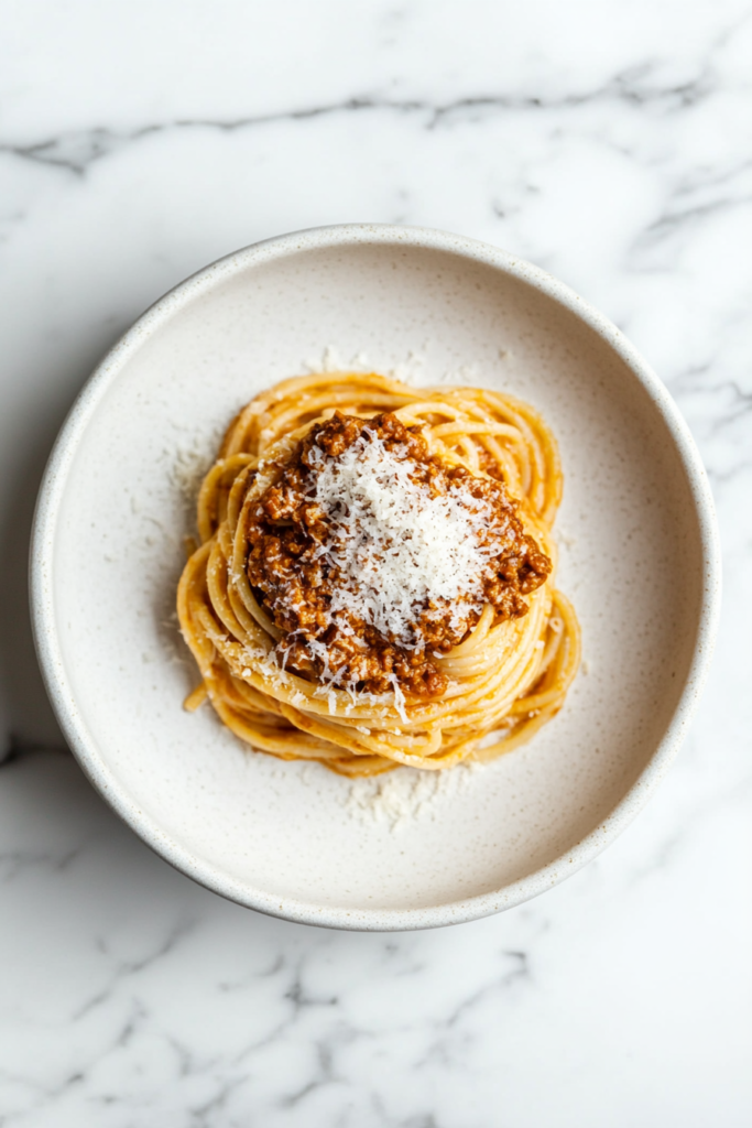 This image shows crockpot bolognese pasta served on a white round plate, topped with chopped meat and shredded cheese.