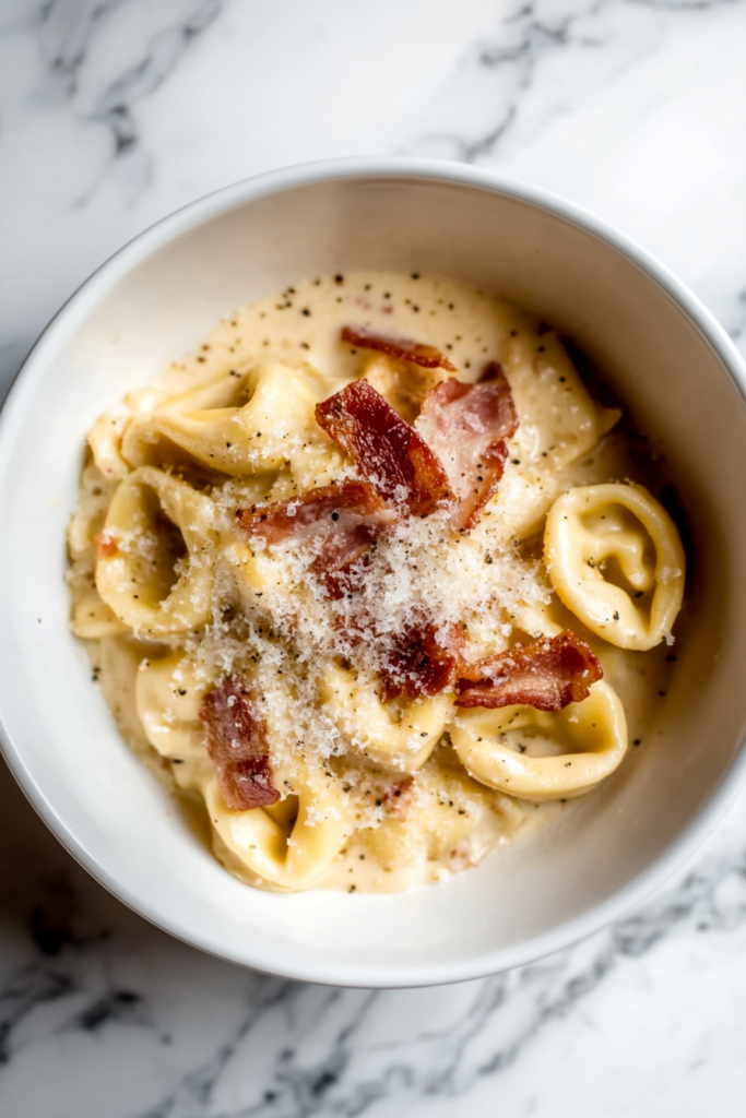 This image shows creamy tortellini pasta carbonara served in a white bowl, topped with bacon bits and shredded cheese.