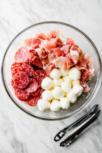 This image shows slices of Italian cured meats and cubes of cheese being added to a salad bowl, creating a hearty and keto-friendly base for the antipasto salad.