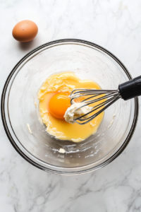 This image shows an egg being cracked and added to the creamed butter and sugar mixture in a bowl.