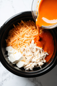 This image shows chunk chicken breast, Buffalo Wing Sauce, ranch dressing, shredded Colby Jack cheese, and softened cream cheese being added to the crockpot as the initial step for preparing Buffalo Chicken Dip.