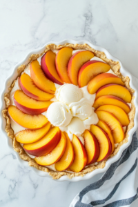 This image shows a layer of cinnamon-coated peach slices being arranged evenly over the softened ice cream layer in the pie crust.