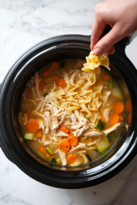 This image shows cooked egg noodles being stirred into the Crockpot Chicken Noodle Soup in the slow cooker.