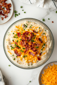 This image shows crispy bacon pieces and shredded cheese being added to the creamy chicken salad mixture in the bowl, enhancing flavor and texture.