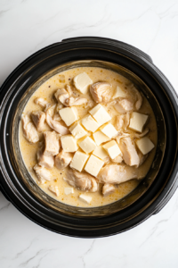 Cubes of cream cheese are scattered over the chicken breasts and cream of chicken soup in the Crockpot.