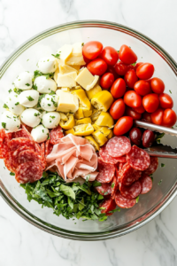 This image shows chopped fresh vegetables, including bell peppers, cherry tomatoes, and olives, being added to the bowl, bringing color and crunch to the antipasto salad.