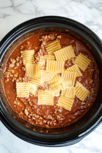 This image shows uncooked lasagna noodles being carefully placed in the crockpot as the first step in layering the lasagna.