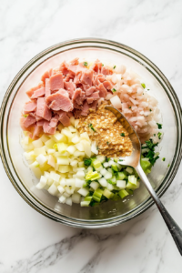 This image shows mayonnaise, Dijon mustard, chopped celery, and garlic powder being added to a bowl of finely chopped ham, preparing the creamy base for the keto deviled ham salad.