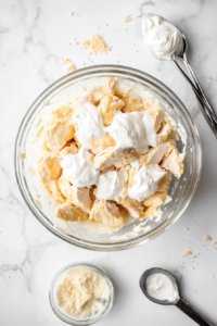 This image shows dollops of mayonnaise and sour cream being added to the shredded chicken in a mixing bowl to create a rich and creamy base for the salad.