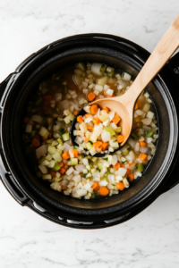 This image shows sautéed vegetables being transferred from the pan into the slow cooker, ready to combine with other ingredients for the soup.