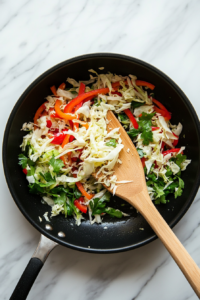 This image shows shredded cabbage being added to the skillet with sautéed bell pepper and scallions, combining to create the base for keto cabbage rice.