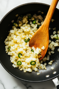 This image shows captures a variety of vibrant spices, including cumin, red pepper flakes, and chili powder, being added to a sautéed onion and jalapeño mixture to enhance the queso dip's flavor.