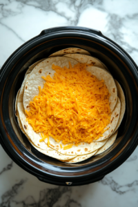 This image shows the final layer of tortillas being topped with a generous amount of shredded cheese in the slow cooker, completing the Crockpot Enchiladas before cooking.