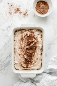 This image shows an even layer of cinnamon-sugar being sprinkled over the first half of the batter in the loaf pan, creating the first layer of sweet and spicy flavor for the bread.