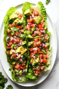 This image shows keto chicken taco lettuce wraps being topped with slices of avocado, diced tomatoes, and fresh cilantro, adding vibrant flavors and textures before serving.