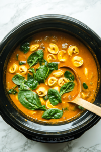 This image shows fresh spinach and tortellini being added to the creamy soup in the crockpot, enhancing the dish's flavor and texture.