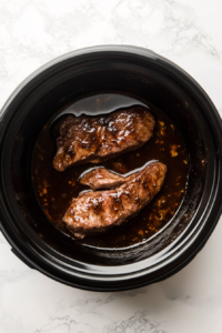 This image shows water, wine, and soy sauce being poured into the slow cooker, combining with the pork and onion soup mix to create a flavorful cooking liquid.