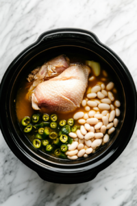 This image shows ingredients like white beans, green chiles, cumin, oregano, and cayenne pepper being added to a slow cooker with chicken to create the Crockpot White Chicken Chili.