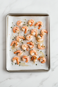 This image shows shrimp being evenly spread on a baking sheet, seasoned and ready to be baked in the oven.