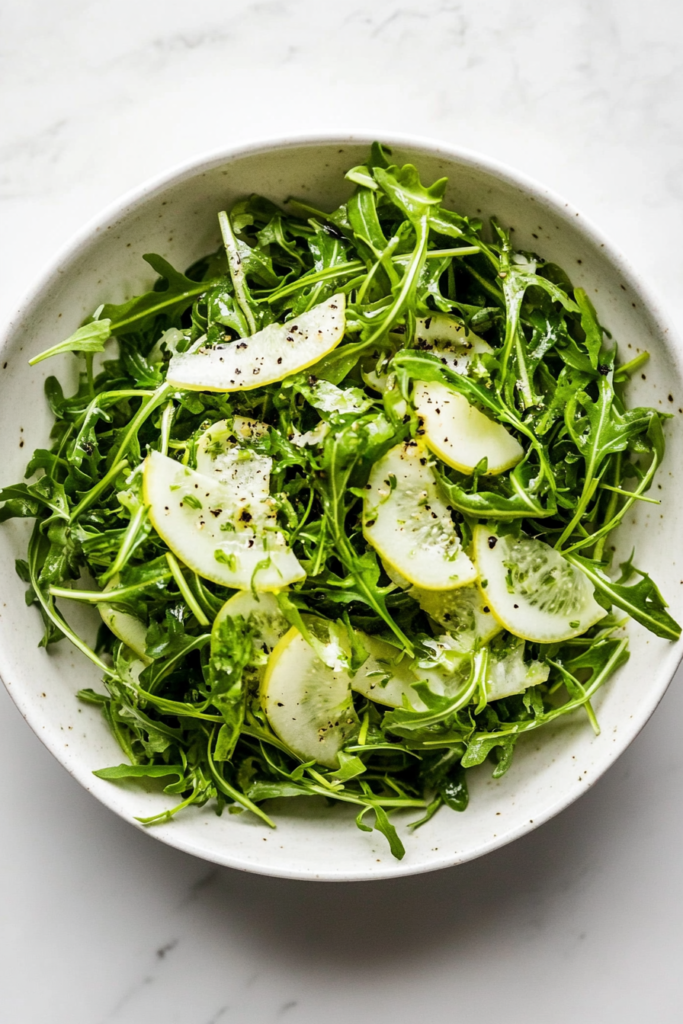 This image shows a white bowl of arugula and thinly sliced fennel, tossed with a zesty dressing made with extra-virgin olive oil, lemon zest, lemon juice, salt, and freshly ground pepper.