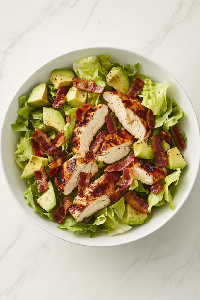 This image shows a vibrant chopped salad in a white bowl, featuring crispy bacon, grilled chicken, and a creamy dressing made with blue cheese, white wine vinegar, water, and pepper, blended to perfection.