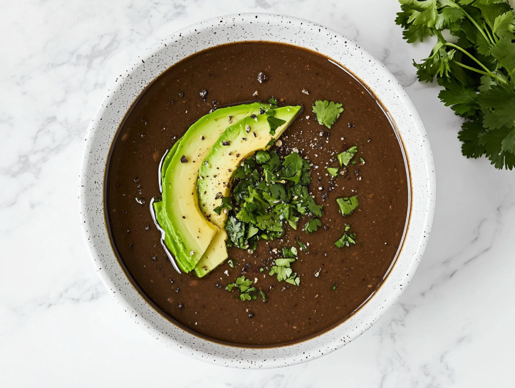 This image shows a hearty black bean soup served in a white bowl, topped with fresh tomato salsa, sliced avocado, and a sprinkle of fresh cilantro for a vibrant and flavorful finish.