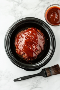 This image shows a brush applying a generous layer of tangy BBQ sauce onto the meatloaf inside the slow cooker, adding flavor and moisture.