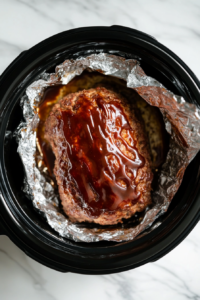 This image shows the fully cooked meatloaf being lifted out of the slow cooker using the aluminum foil lining, showcasing its tender and juicy texture.