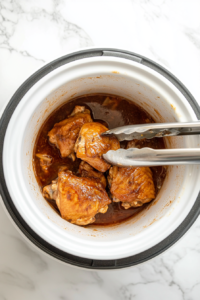 This image shows a pair of tongs lifting a cooked chicken thigh from the slow cooker, demonstrating its juicy tenderness and flavorful glaze.