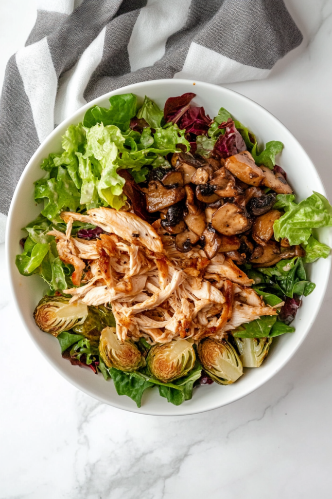 This image shows a hearty salad in a white bowl with grilled chicken, shredded Brussels sprouts, and sautéed mushrooms, dressed with olive oil, red wine vinegar, minced shallot, Dijon mustard, chopped thyme, and ground pepper.