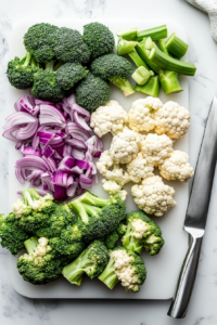 this image shows Fresh broccoli, cauliflower florets, and red onion diced on a cutting board.