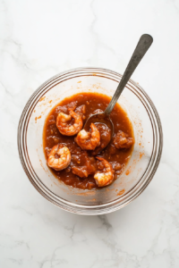 This image shows large, cooked shrimp being coated evenly in a tangy barbecue sauce mixture, ready to be wrapped in bacon for air frying.