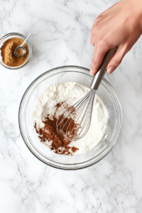 This image shows a bowl of flour, baking soda, salt, and spices being whisked together to form the dry ingredients.