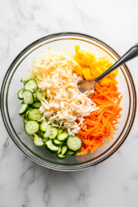 This image shows sliced cucumbers, shredded crab sticks, and diced mango being mixed together in a bowl, preparing the base of the kani salad.