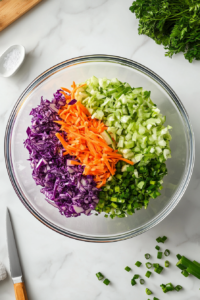 this image shows a large mixing bowl filled with shredded green cabbage, red cabbage, carrots, and sliced green onions, ready to be combined for a vibrant keto coleslaw.