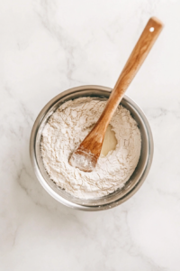 This image depicts flour being stirred into melted coconut oil in a pan, forming a golden roux, the essential base for creamy vegan béchamel sauce.