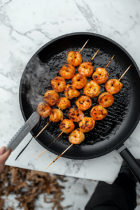 Marinated shrimp skewers being grilled over an open flame, showing char marks and smoky flavor development.