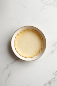 This image shows a golden-brown pie crust cooling in a white pie dish, ready to be filled with peach-flavored cream and fresh peaches for a delicious dessert.