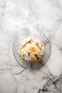 This image shows the golden-brown loaf of no-knead Dutch oven bread resting on a wire rack, cooling to room temperature to ensure the interior sets properly.