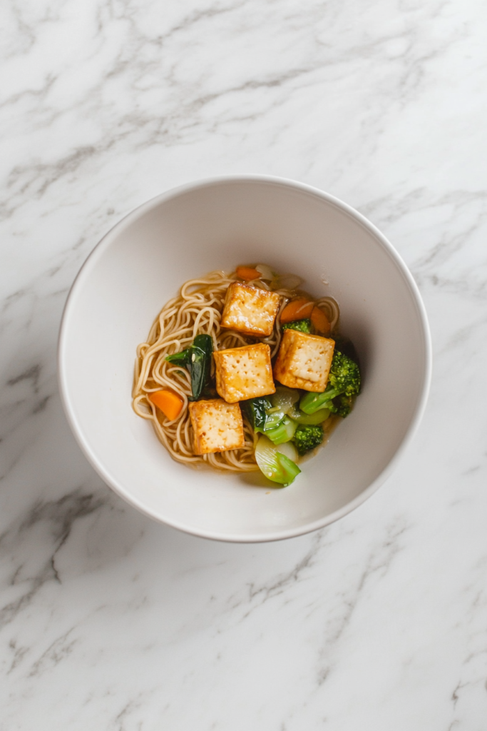 This image shows a white round serving dish filled with crispy egg noodles, topped with shredded carrots, cabbage, fresh cilantro, mint, and crispy tofu, creating a colorful and flavorful vegetarian meal.