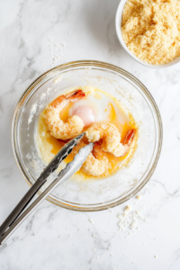 This image shows a plump shrimp being dipped into a bowl of egg wash, ensuring it’s fully coated and ready for the cornmeal mixture.