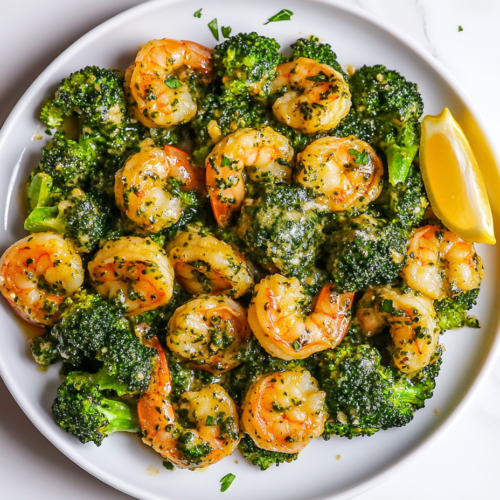 This image shows a beautifully plated garlic butter shrimp broccoli skillet on a white round plate. The dish features tender shrimp and vibrant green broccoli, topped with a squeeze of fresh lemon juice, parsley, lemon slices, and a sprinkle of crushed red chili pepper.