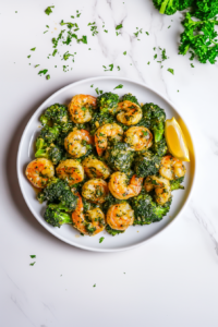 This image shows the finished garlic butter shrimp broccoli skillet being garnished with fresh parsley, lemon slices, and crushed chili flakes, with final heating to bring all the flavors together.