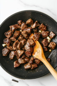 This image shows a hand sprinkling freshly chopped parsley over golden brown steak bites, adding a vibrant green touch to the dish.