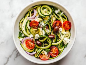 This image shows a vibrant Greek zucchini salad served in a white bowl, featuring zucchini noodles, kalamata olives, and a tangy dressing made with olive oil, vinegar, garlic, and oregano, topped with crumbled feta cheese for a delicious and healthy meal.