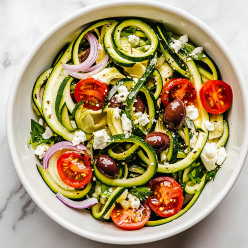 This image shows a vibrant Greek zucchini salad served in a white bowl, featuring zucchini noodles, kalamata olives, and a tangy dressing made with olive oil, vinegar, garlic, and oregano, topped with crumbled feta cheese for a delicious and healthy meal.