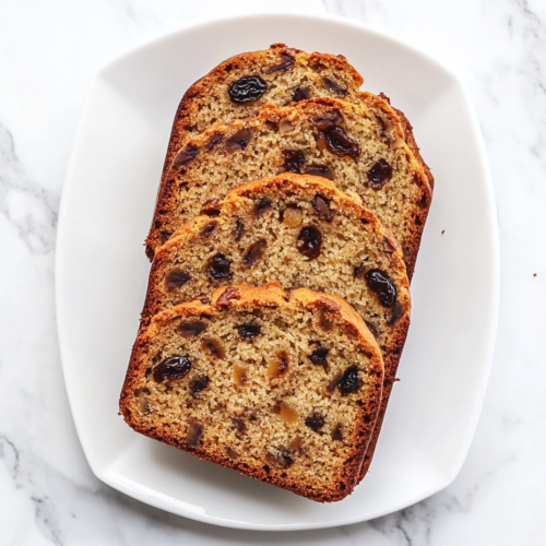 This image shows a loaf of Jamaican banana bread on a white plate, garnished with visible pecans and raisins, with slices cut and arranged around it.