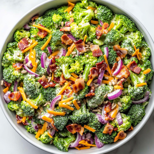 This image shows a white bowl filled with keto broccoli salad, featuring bite-sized broccoli florets, shredded cheese, chopped onions, walnuts, sunflower seeds, crumbled bacon, and creamy dressing evenly mixed for a delicious low-carb dish.