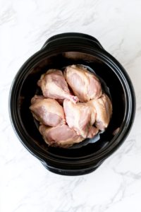 Raw chicken breasts are placed at the bottom of a slow cooker, forming the base layer for chicken fajitas.